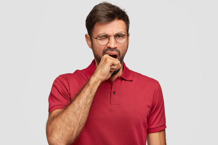 A man with a beard and glasses wearing a maroon polo shirt, yawning and covering his mouth with his hand against a plain light background.