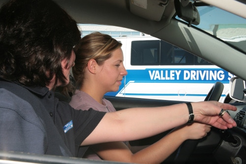 Female student driver receiving hands-on instruction from a driving Instructor inside a car, with a Valley Driving School van in the background.