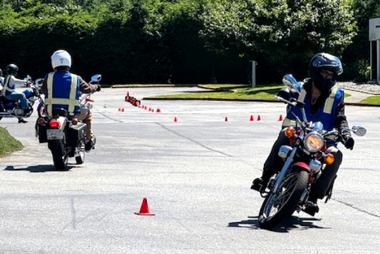 Several motorcyclists wearing helmets and high-visibility blue safety vests practice maneuvers around orange traffic cones on an outdoor asphalt training lot.