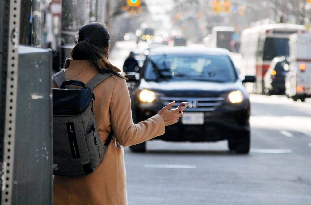 Rear view of a woman with a backpack holding a smartphone to book a ride-hailing service as a black SUV approaches in the city.