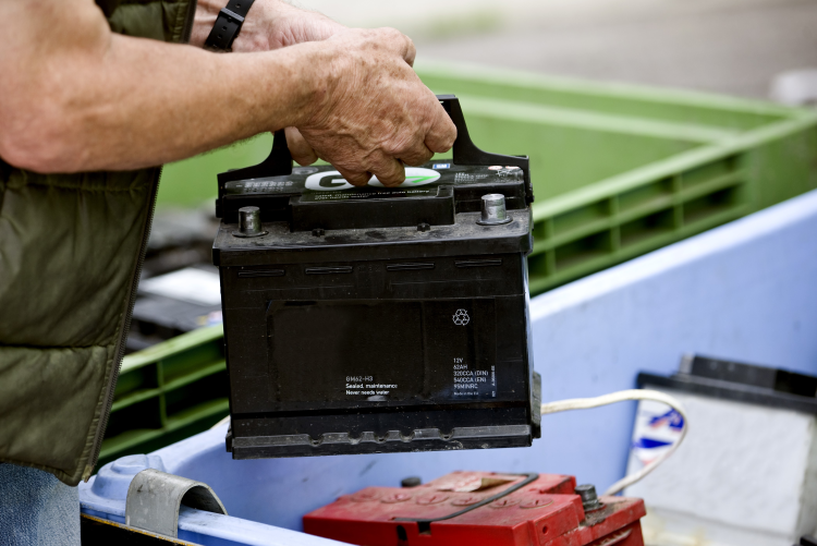A person carrying a black car battery by its handle to place it into a blue recycling bin containing other used batteries.