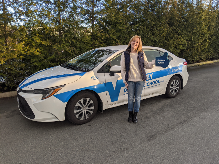 A happy student driver holding a blue "#ipassed" foam thumbs-up sign while standing next to a branded Valley Driving School training car.