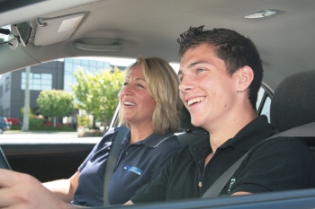 A smiling female driving instructor and a young male student during a positive behind-the-wheel driving lesson.