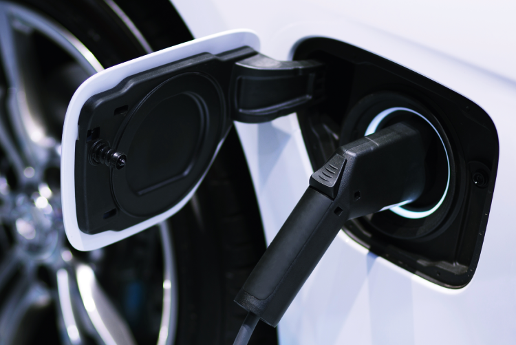 A close-up of a black electric vehicle charging connector plugged into the open charging port of a white car, featuring a subtle blue glow around the plug.