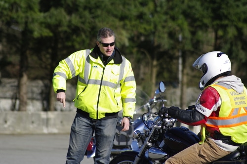 A valley driving school motorcycle instructor in a high-visibility jacket points toward the ground while coaching a student on a motorcycle.