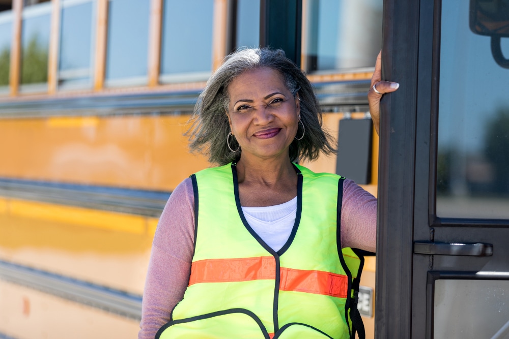 Female school bus driver in a high-visibility vest standing by a yellow school bus, smiling.