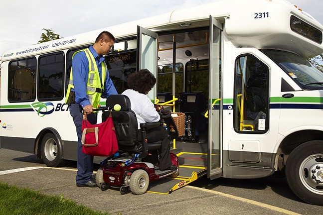 BC Transit operator assisting a passenger in a mobility scooter onto a HandyDART bus using a vehicle ramp.