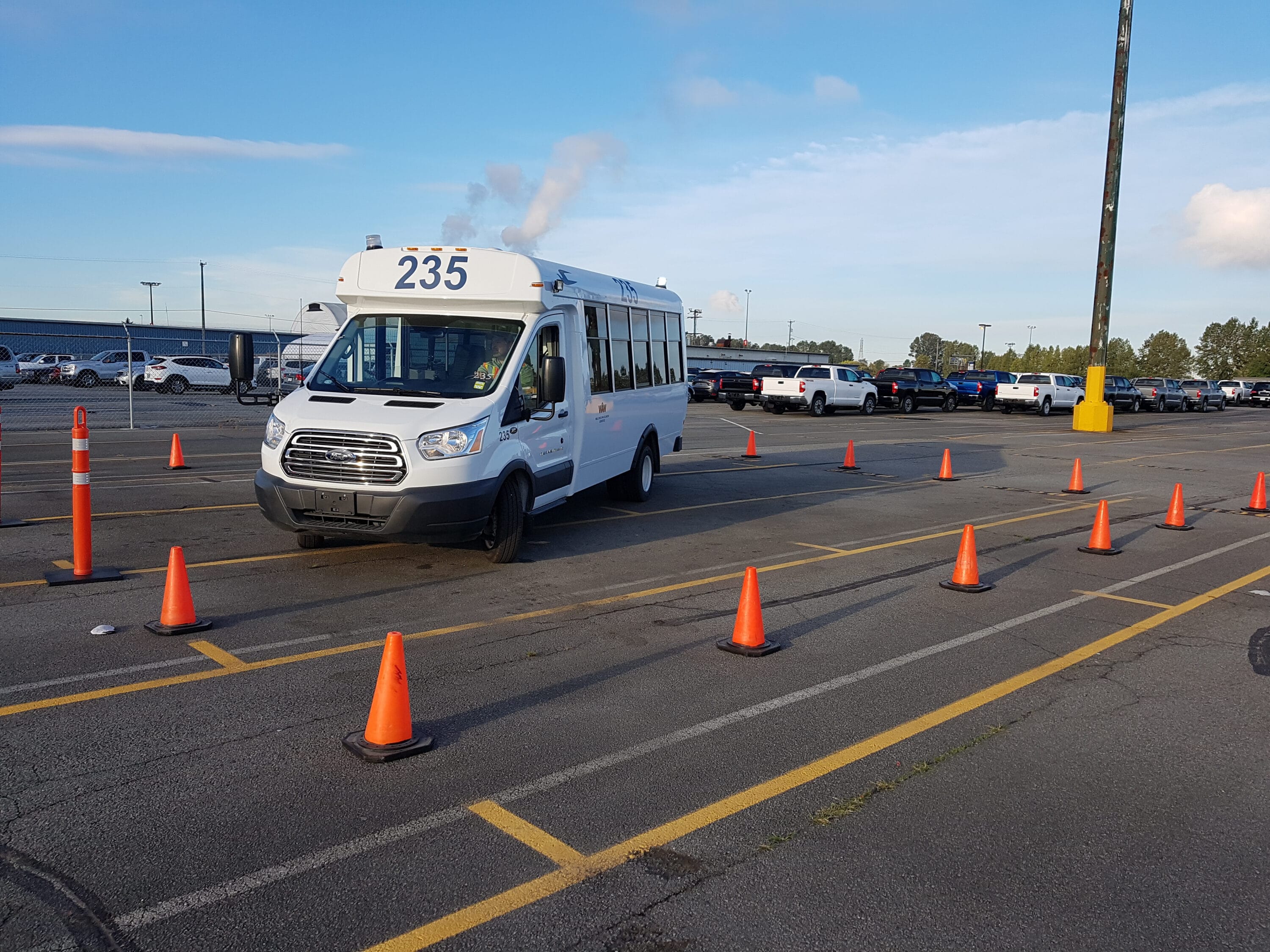 A white shuttle van in a parking lot, maneuvering around orange traffic cones.