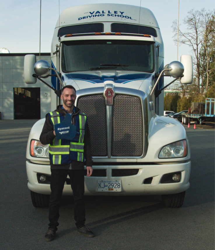 A man wearing a blue safety vest is smiling and standing in front of a large semi truck, holding a sign that says "I passed".