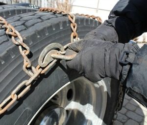 A student driver demonstrating the proper technique for securing snow chains on a tractor-trailer tire during a winter safety course.