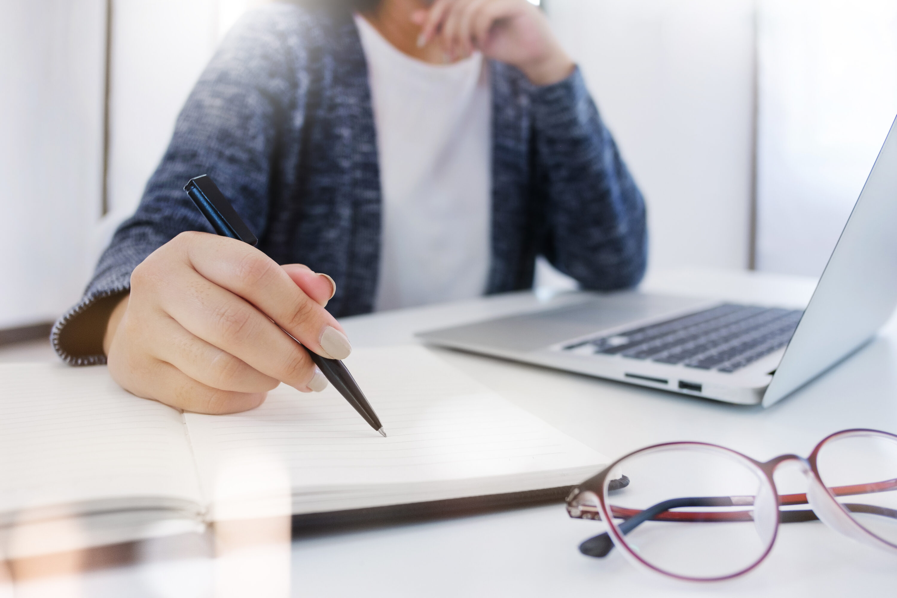 a person sitting at a white desk with a silver laptop.  They are writing in a note book with a pair of glasses beside the notebook.