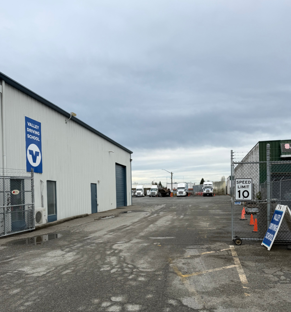 A driveway into a gravel truck yard with multiple semi trucks parked at the rear of the yard. There is a building to the left of the driveway that has a Valley Driving School blue sign on it.