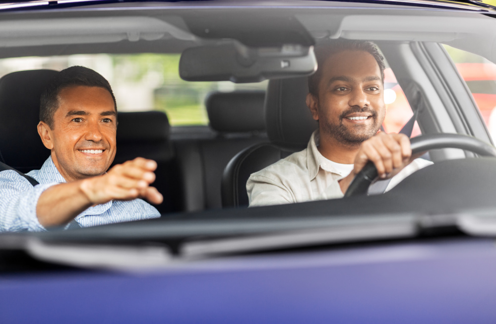 A male driving Instructor is gesturing to a male student who is holding onto the steering wheel. Both men are smiling.
