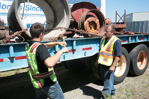 A Valley Driving School instructor supervising a student as they use a winch bar to secure heavy concrete cargo with chains on a flatbed trailer.