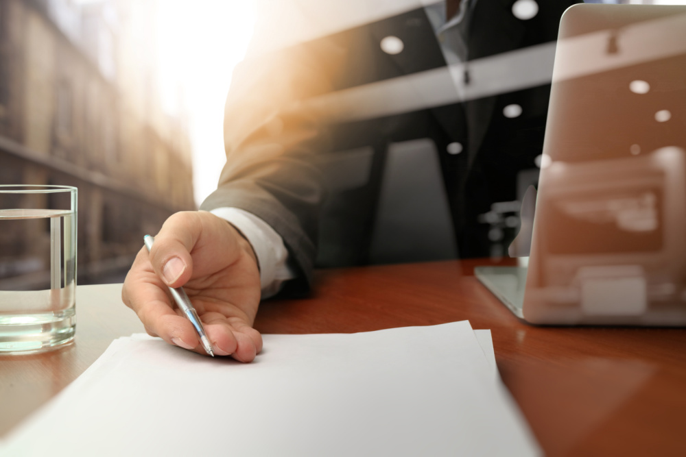 a man sitting across a desk holding a pen and pointing towards a paper contract.