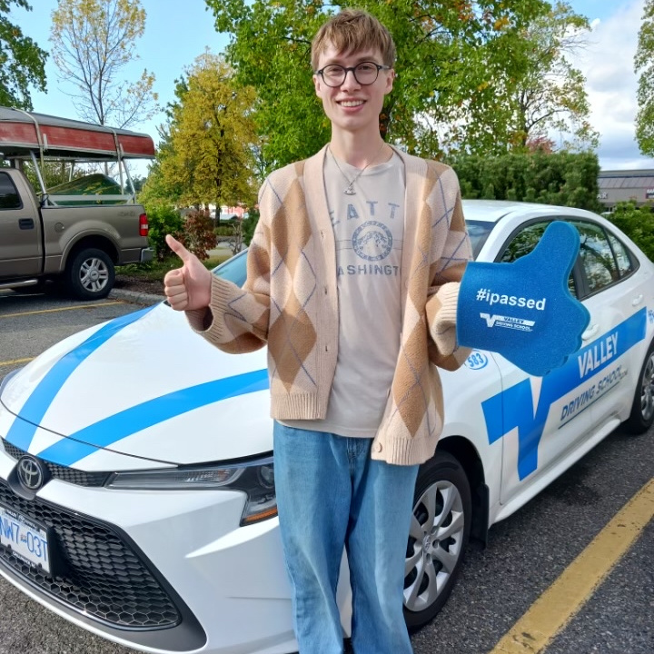 A smiling young man stands with two thumbs up next to a branded white Valley Driving School Toyota Corolla, holding a blue sign that reads "#ipassed" and the "Valley Driving School" logo.
