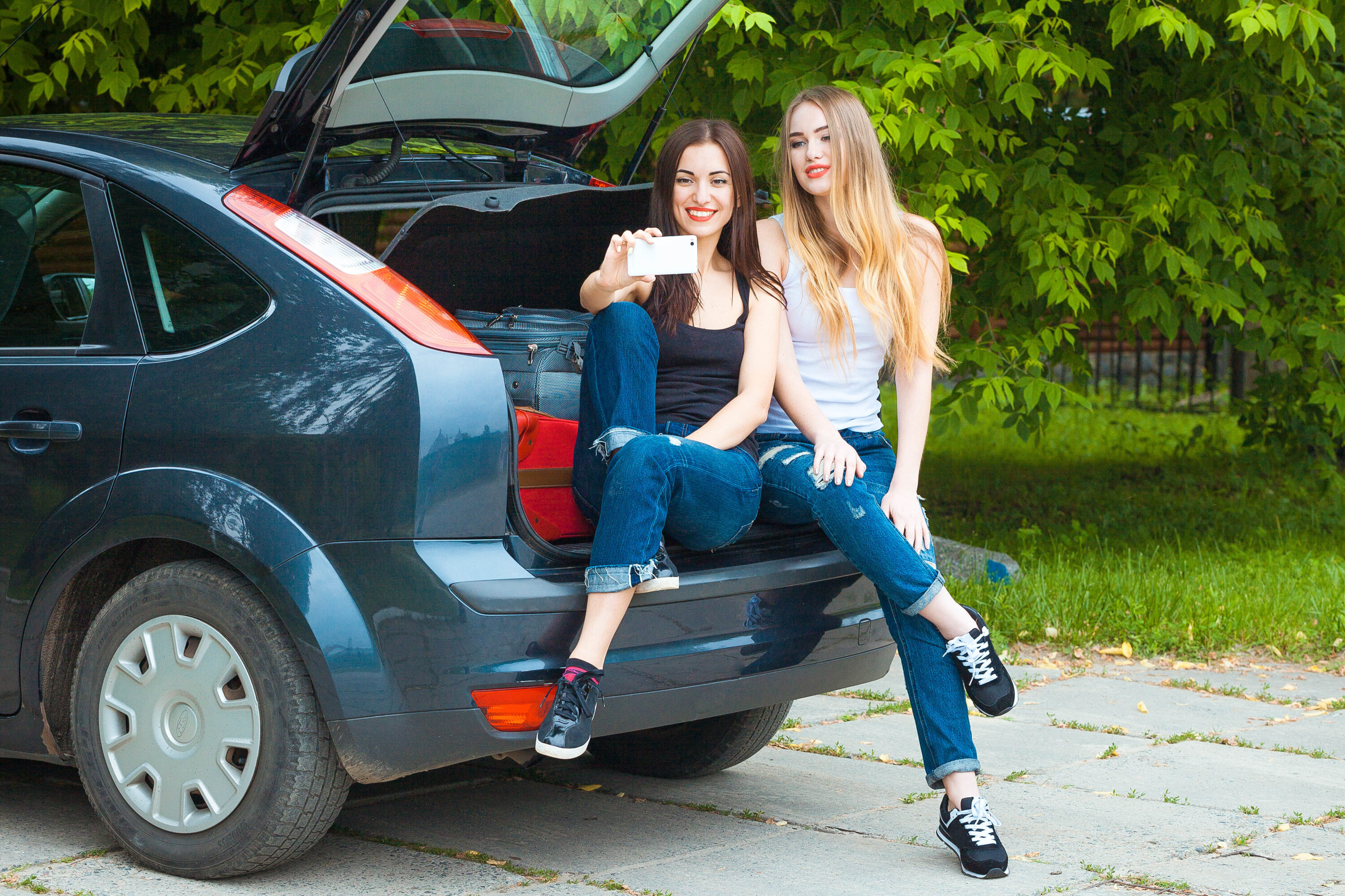 2 young girls taking a selfie while sitting on the back end of a blue car.