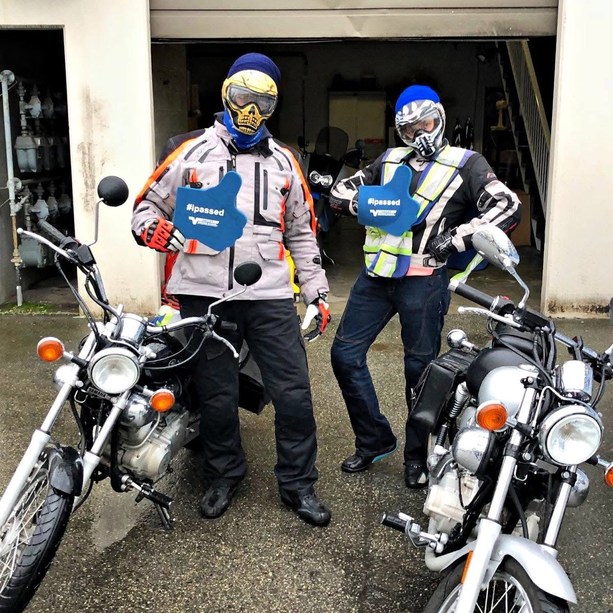 2 motorcycle riders in full gear, standing beside their bikes and holding signs that say "I passed".