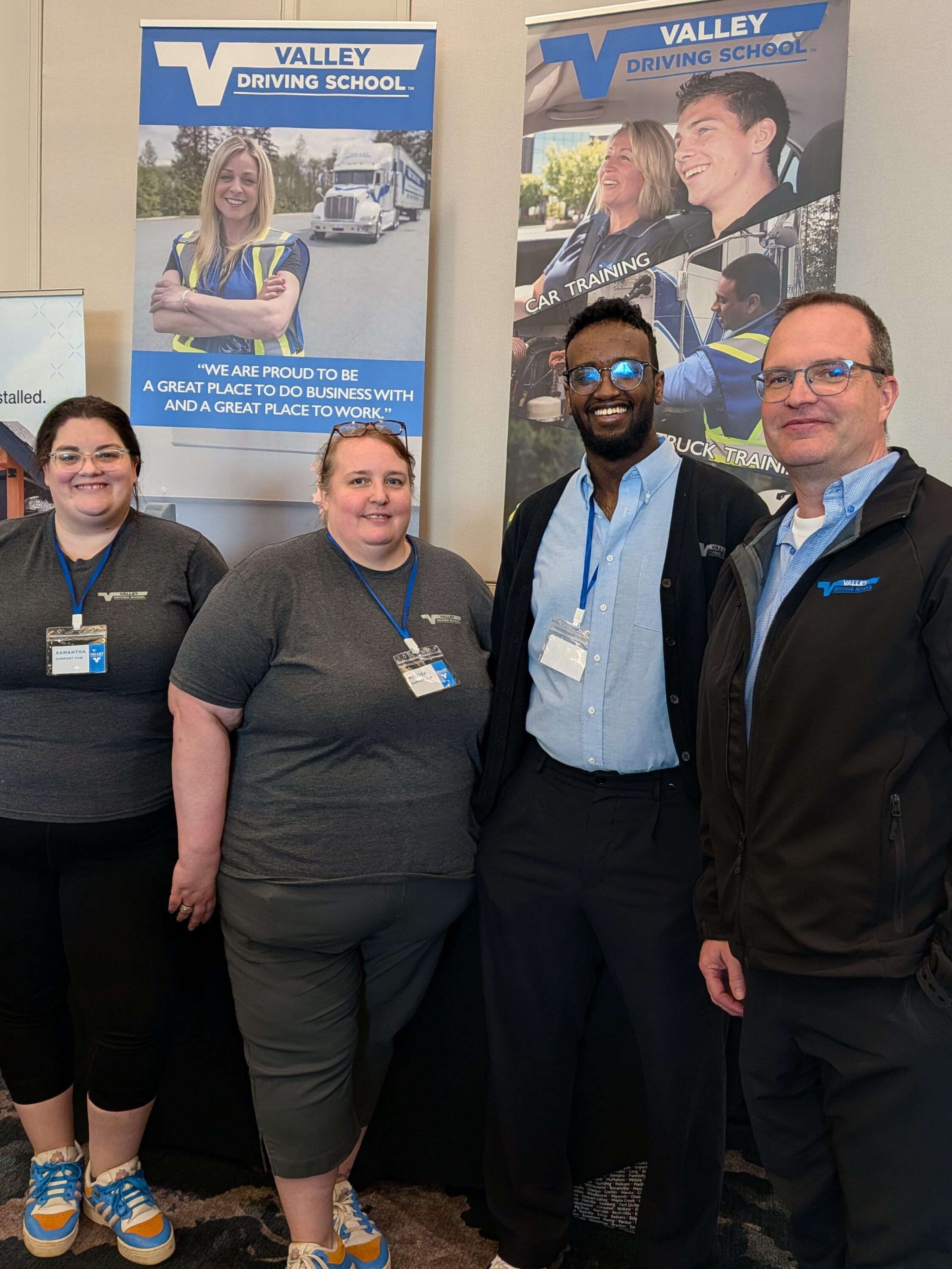 Four Valley Driving School team members smiling and posing together in front of company banners.