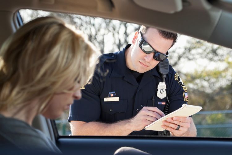 A police officer writing a traffic ticket for a driver seated inside a vehicle.